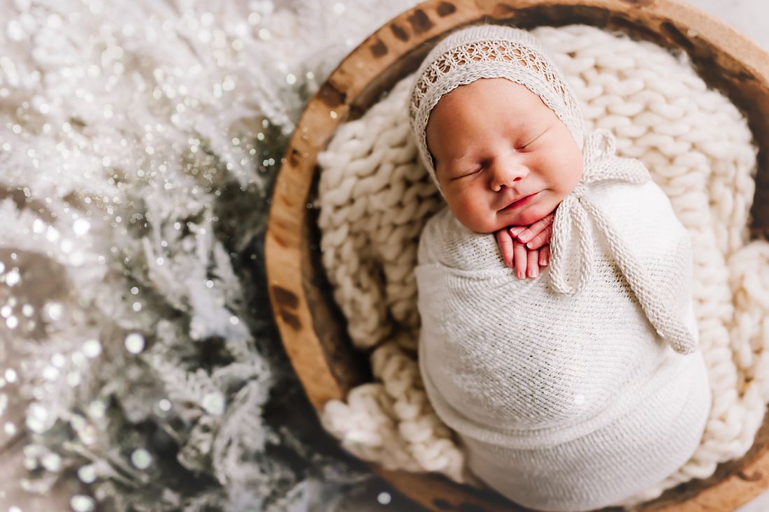 Overhead view of a newborn in a cream knit bonnet and wrap, swaddled and asleep in a round wooden bowl beside frosted greenery